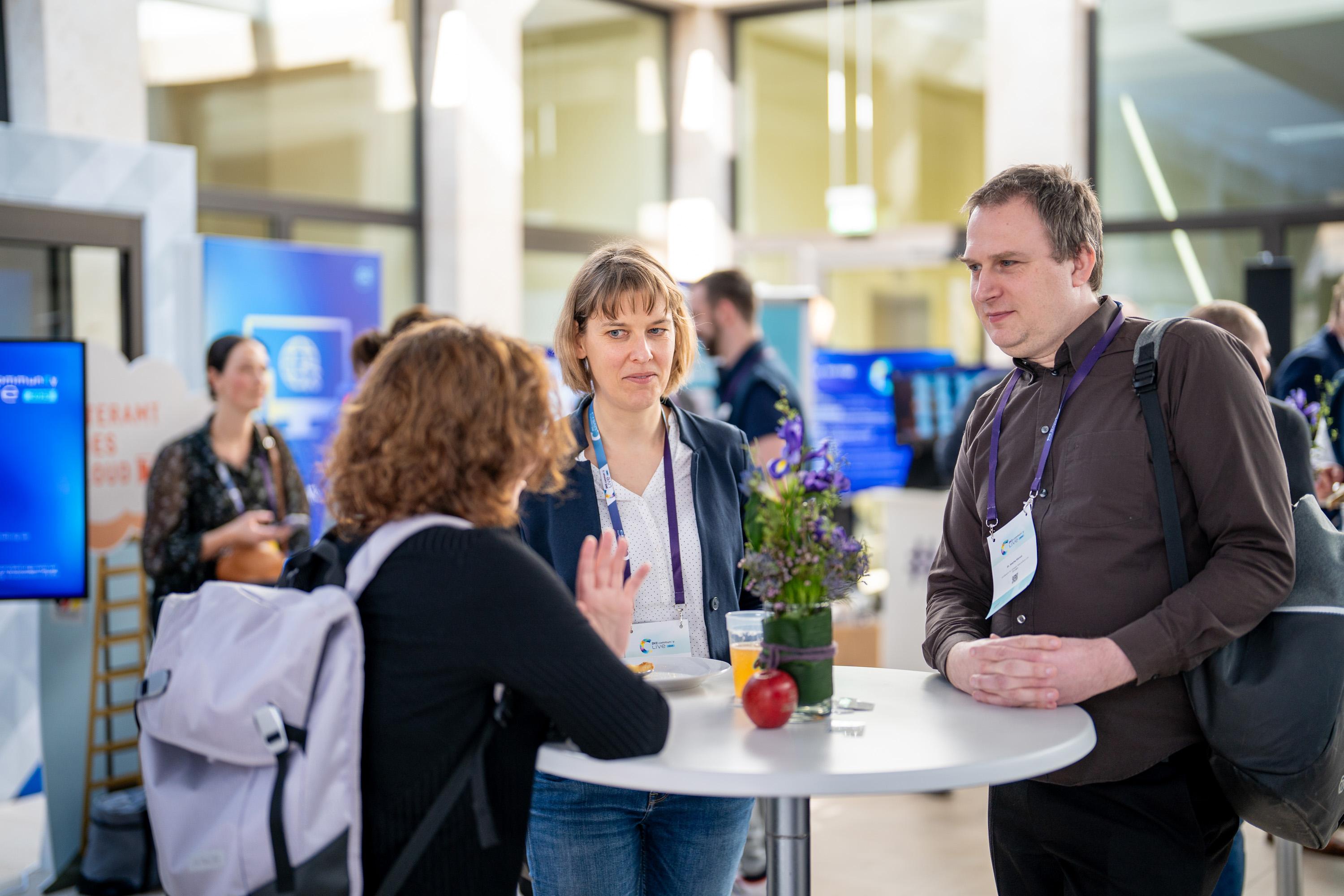 Drei Personen stehen an einem Stehtisch im Atrium, im Hintergrund sind weitere Teilnehmer und Präsentationen zu sehen.