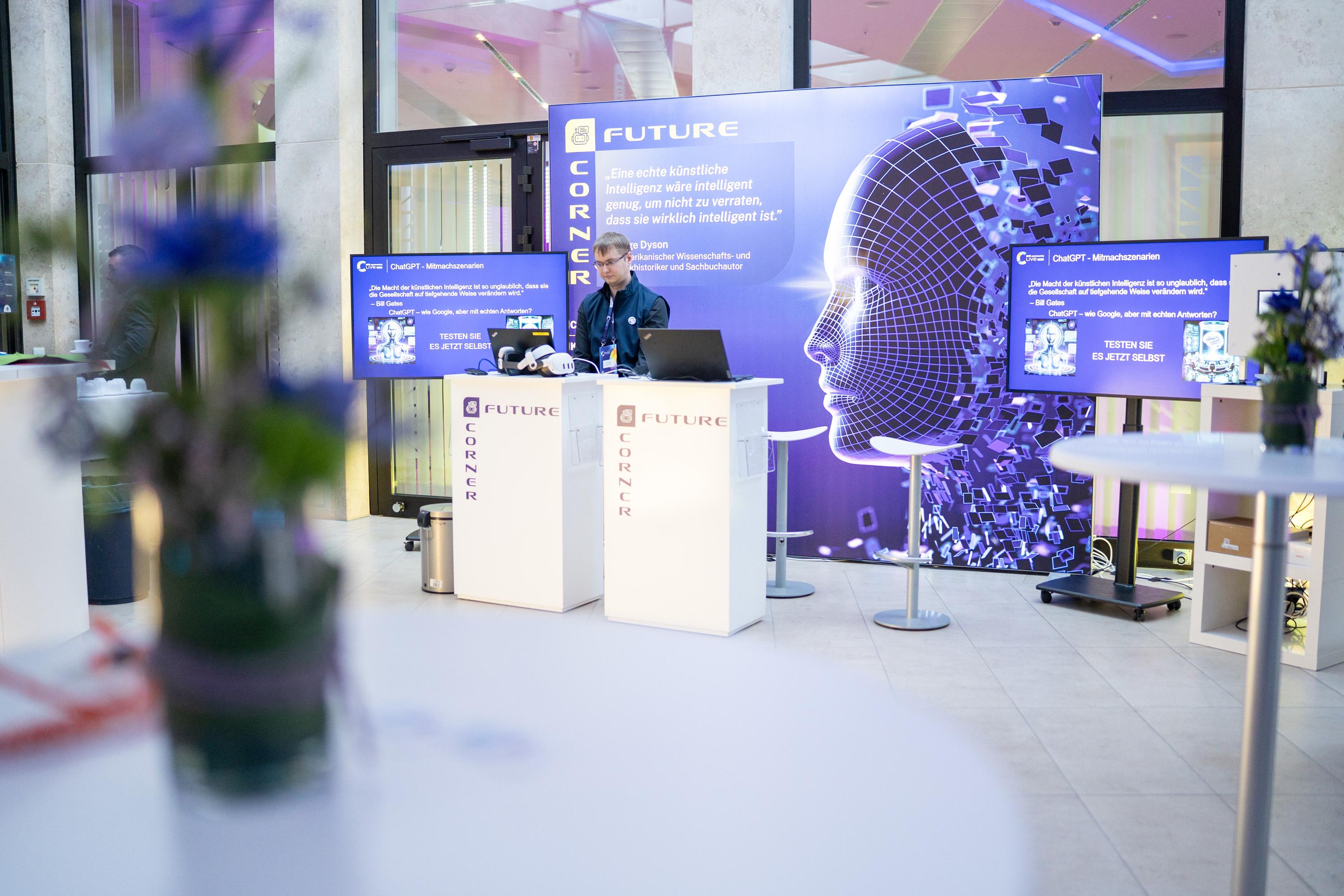 Ein Messestand mit einem Bildschirm, der eine digitale Kopf-Silhouette zeigt. Im Vordergrund stehen Tische mit Blumen. Eine Person steht am Messestand..