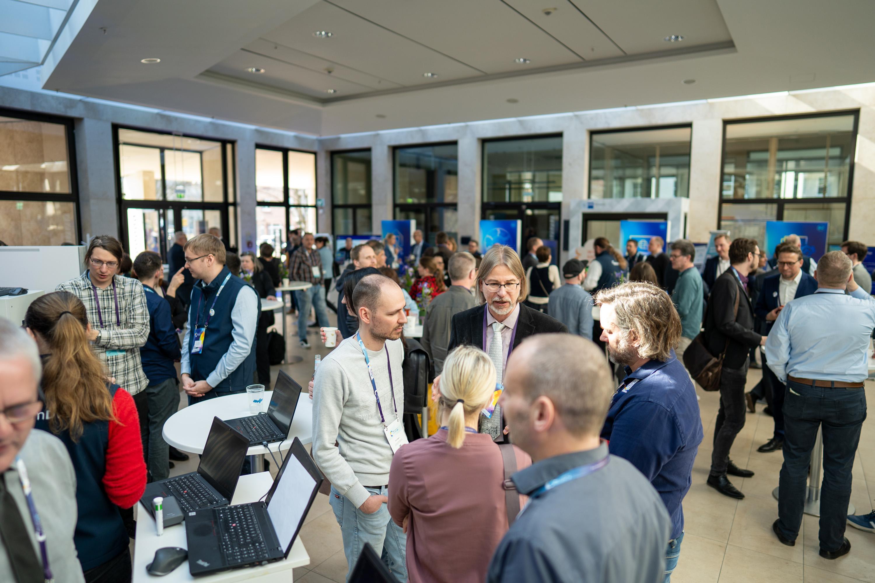 Viele Teilnehmer stehen im Atrium an Stehtischen und den Mesständen und unterhalten sich.