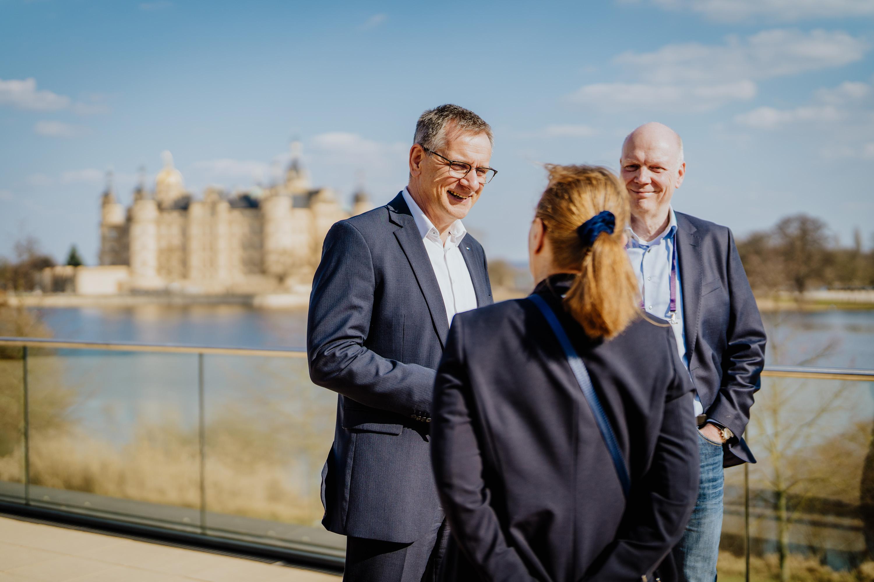 IHK-Geschäftsführer Peter Todt, Uta Knöchel und ein Mann unterhalten sich auf der Dachterrasse der IHK zu Schwerin. Im Hintergrund ist das Schweriner Schloss zu sehen.