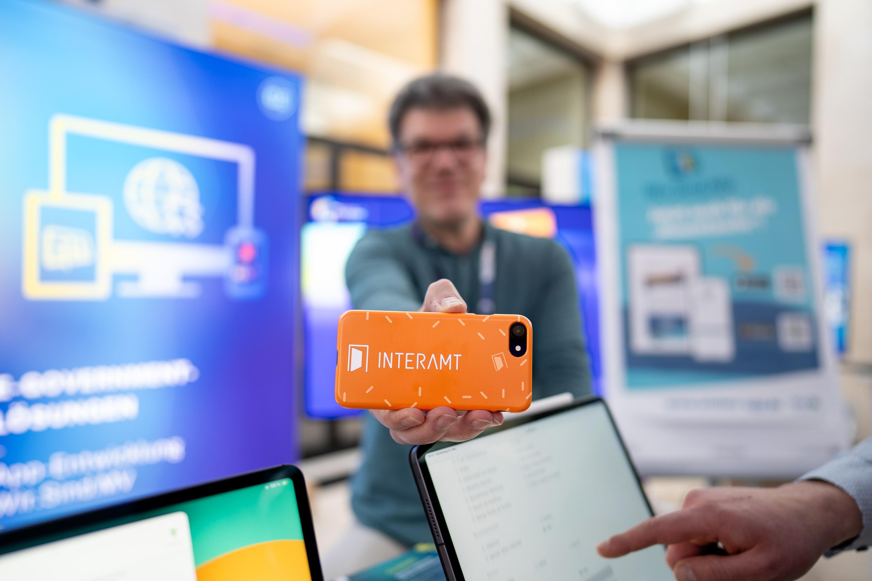 Ein Mann hält ein Handy nach vorne, auf dessen Rückseite ist das Logo von Interamt zu sehen.