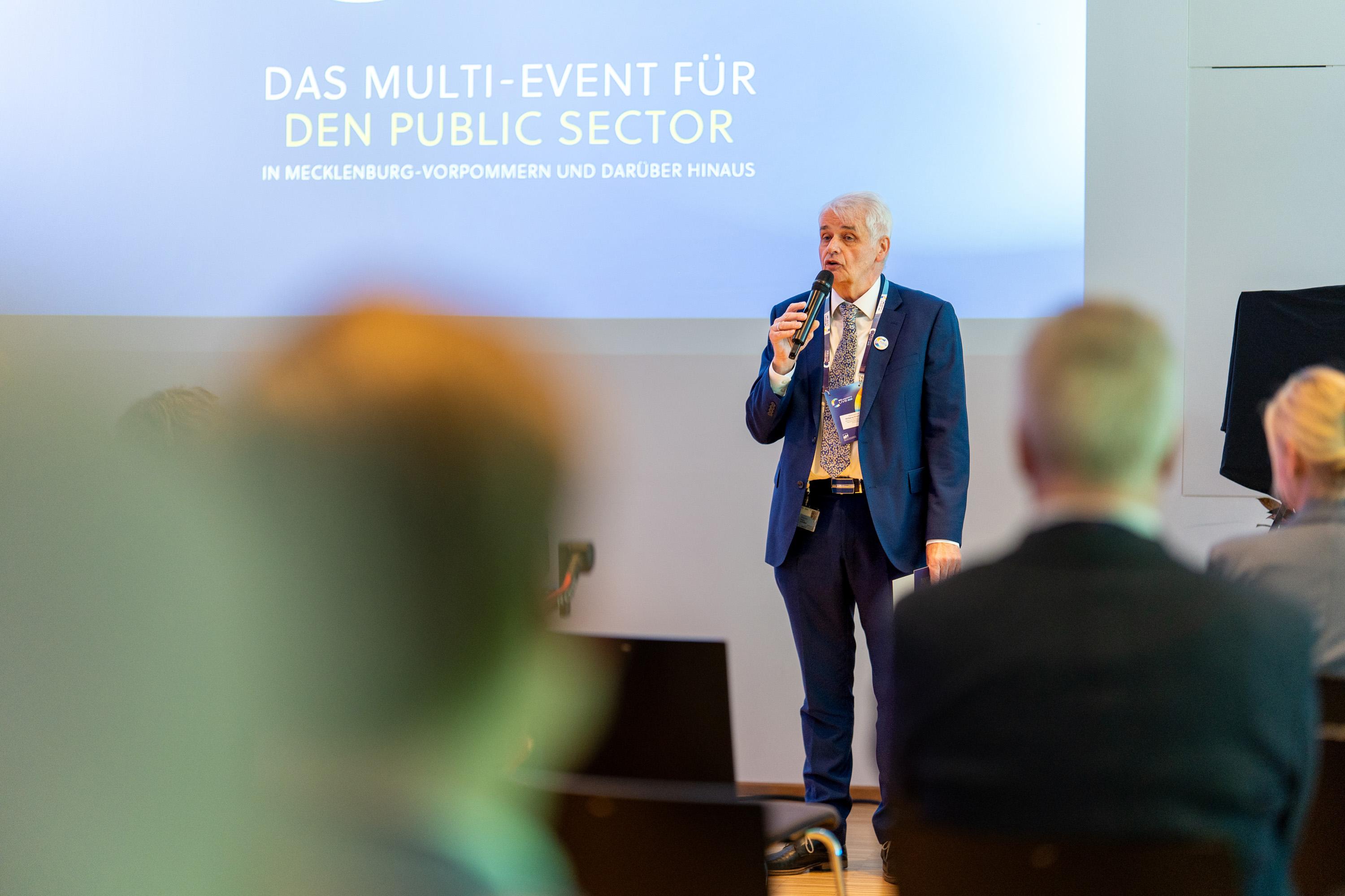 Ein Mann spricht am Mikrofon auf der Bühner, im Hintergrund ein Banner mit dem Eventtitel.