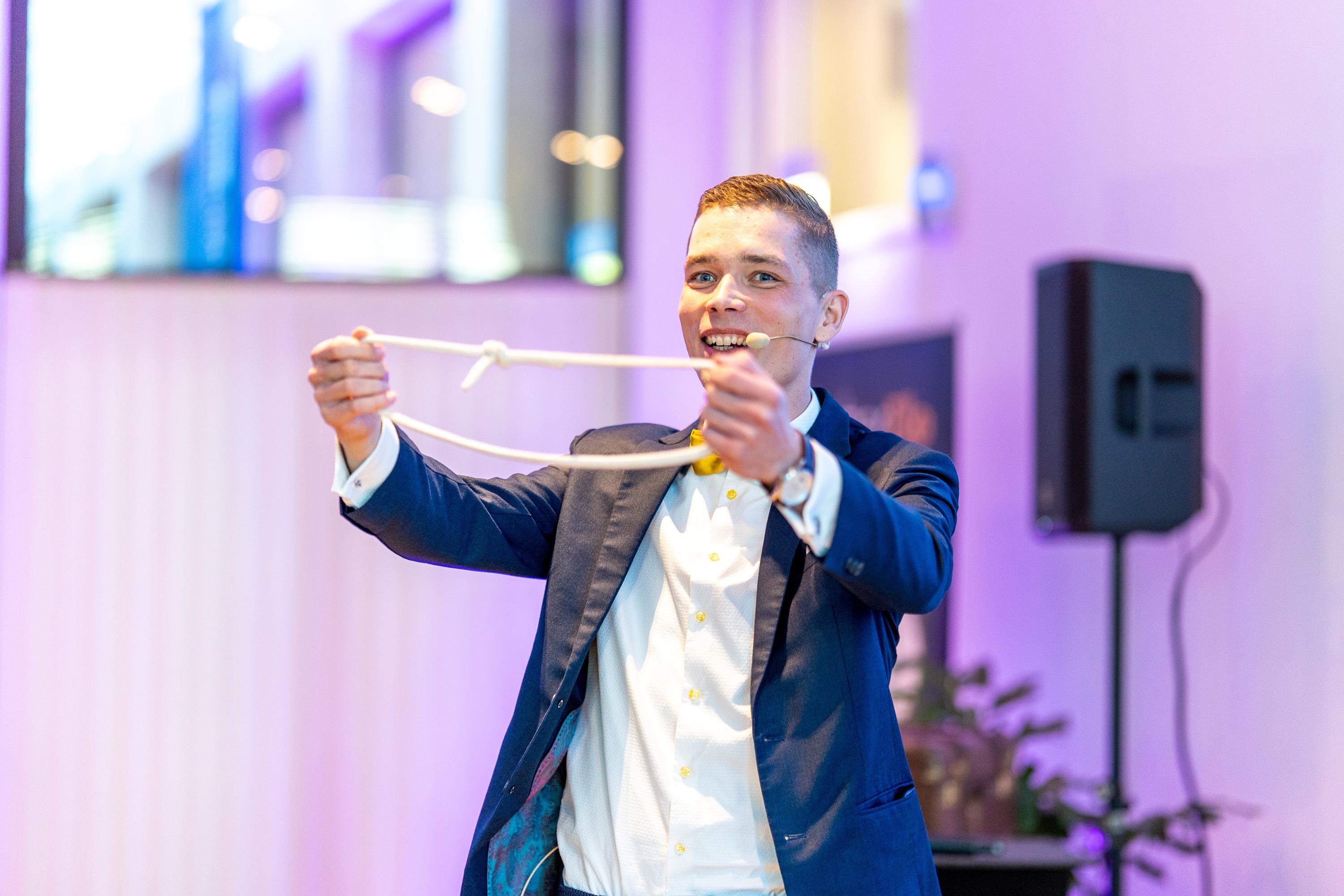 Ein junger Zauberer in einem Anzug mit Fliege, der begeistert ein Seil hält und lächelt. Hintergrund mit unscharfen Lichtern und einem Lautsprecher.