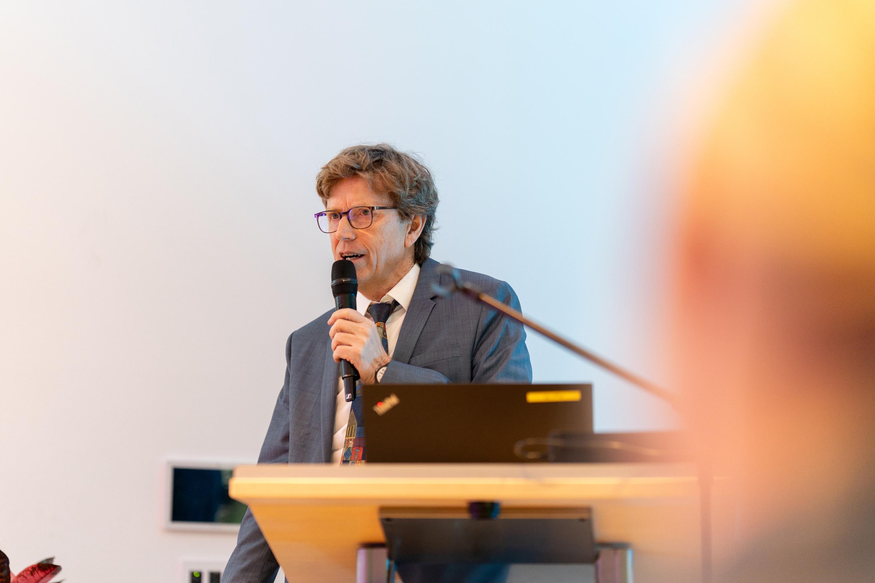 Prof. Dr. Engelbert Lütke Daldrup hält ein Mikrofon und spricht auf einer Bühne im Raum Mecklenburg, im Hintergrund ein Laptop.