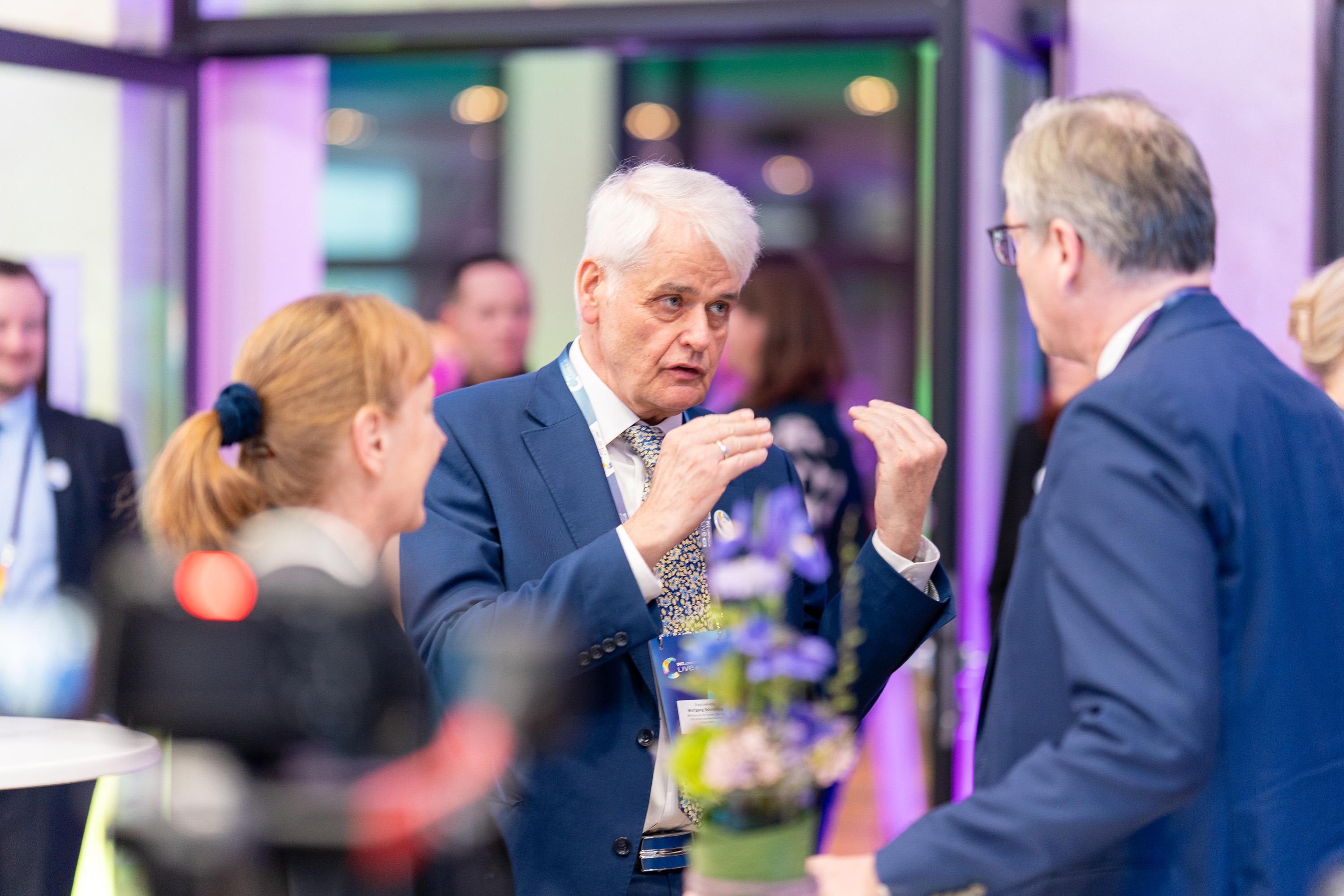 Staaatssekretär Wolfgang Schmülling spricht gestikulierend mit zwei Personen im Atrium, Blumen im Vordergrund.