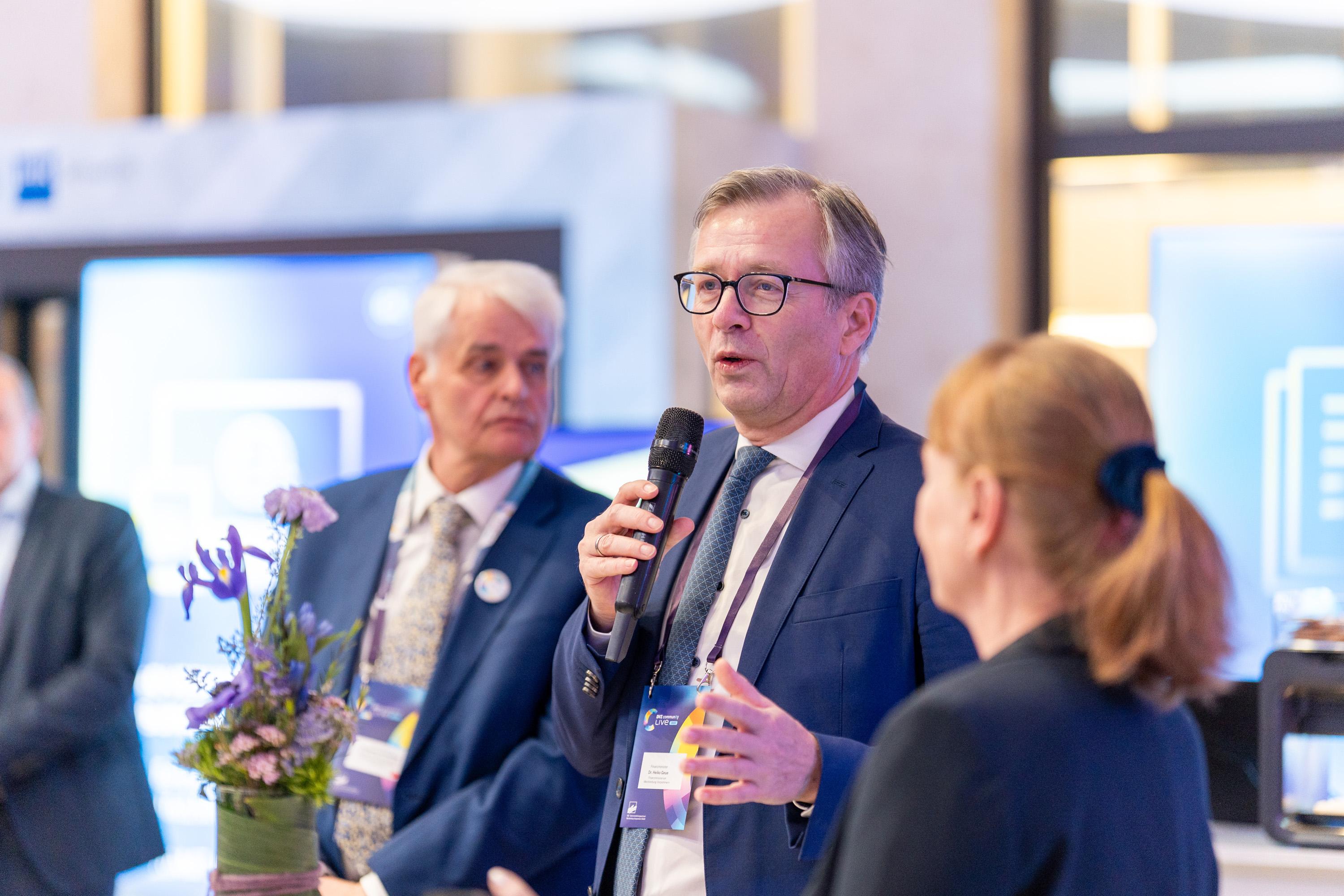 Finanzminister Dr. Heiko Geue spricht am Mikrofon, im Hintergrund zwei Personen und eine Blumendekoration.