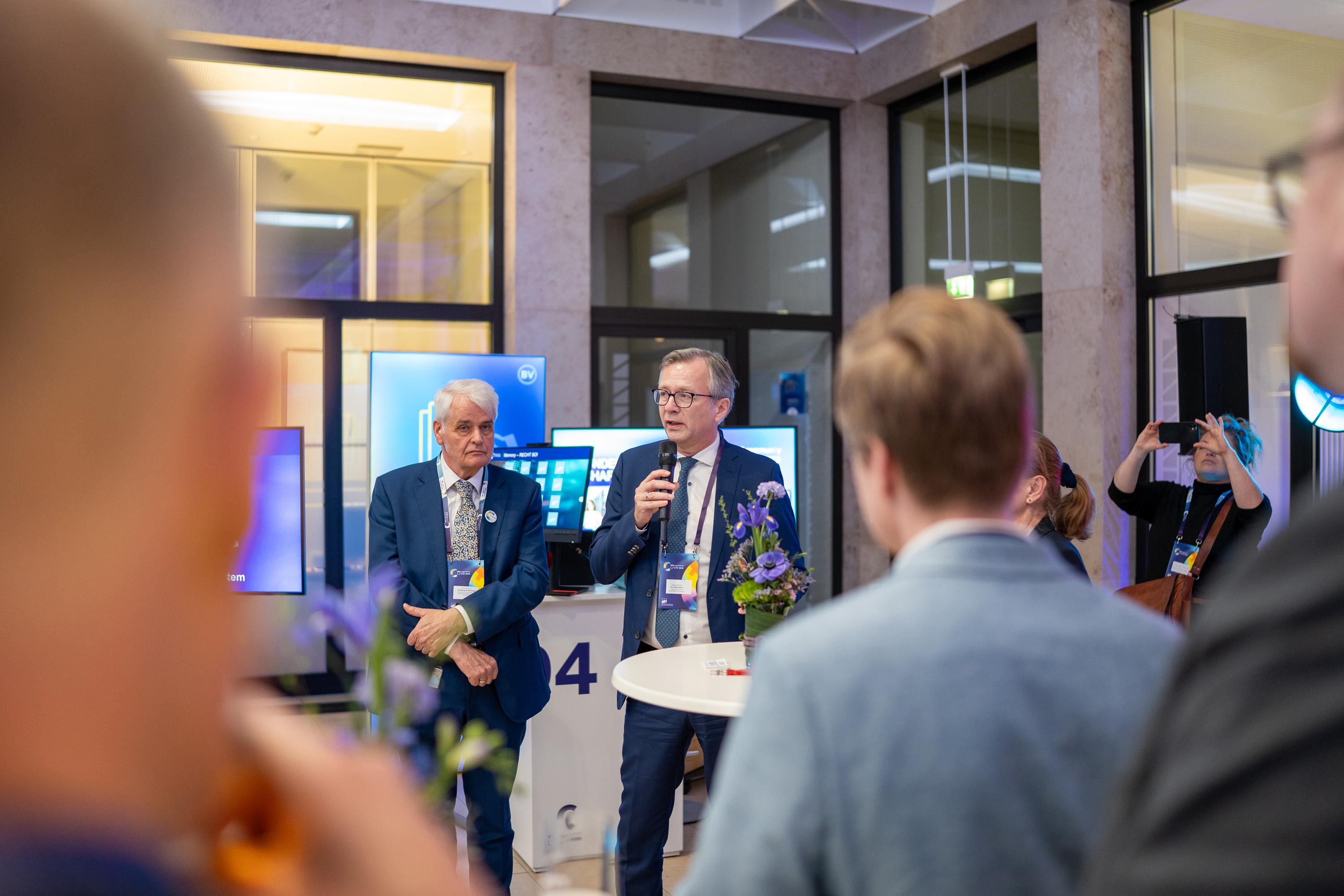 Finanzminister Dr. Heiko Geue und ein Staatssekretär stehen an einem Tisch im Atrium. Im Hintergrund sind weitere Personen und Bildschirme sichtbar.