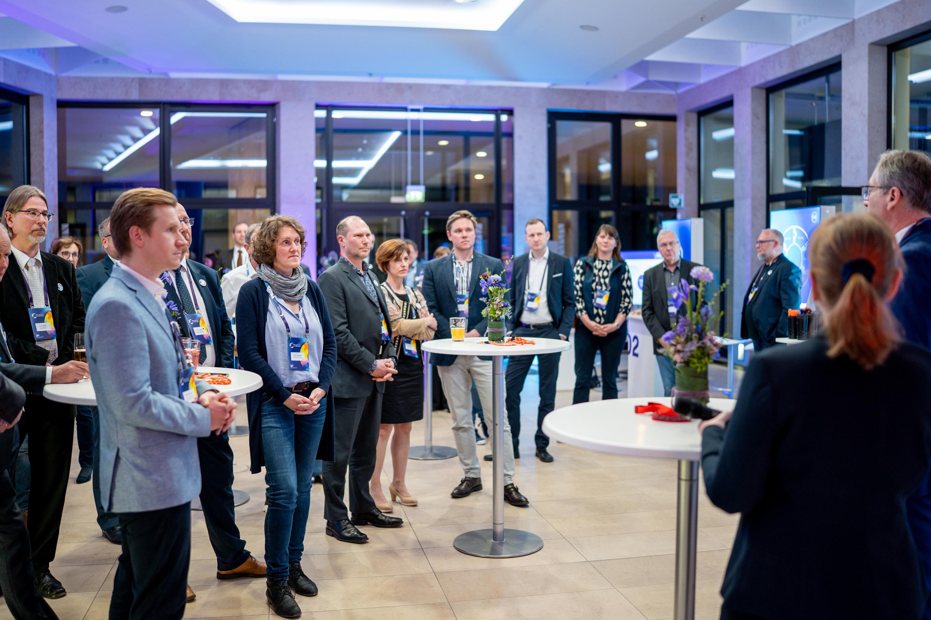 Eine große Gruppe von Teilnehmern steht im Atrium und hört einer Rede zu.