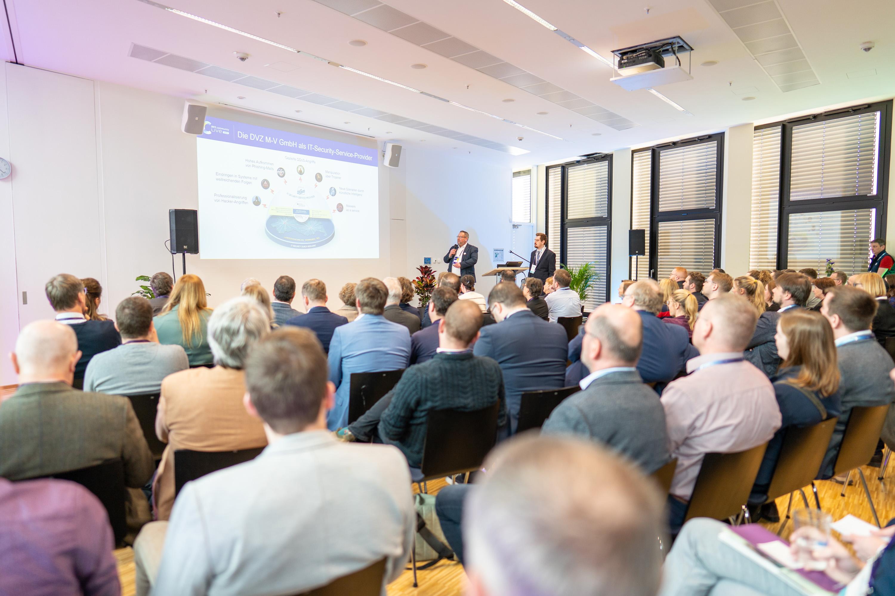 Publikum von hinten zu sehen, vorne präsentieren zwei Speaker