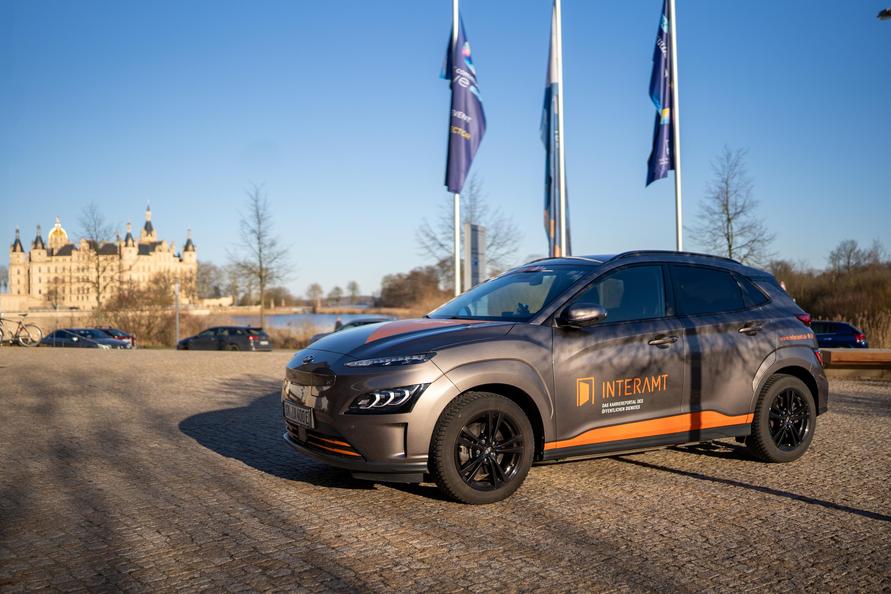 Auto mit dem Logo von Interamt steht auf einem Platz vor dem Schweriner Schloss und Fahnen im Hintergrund.