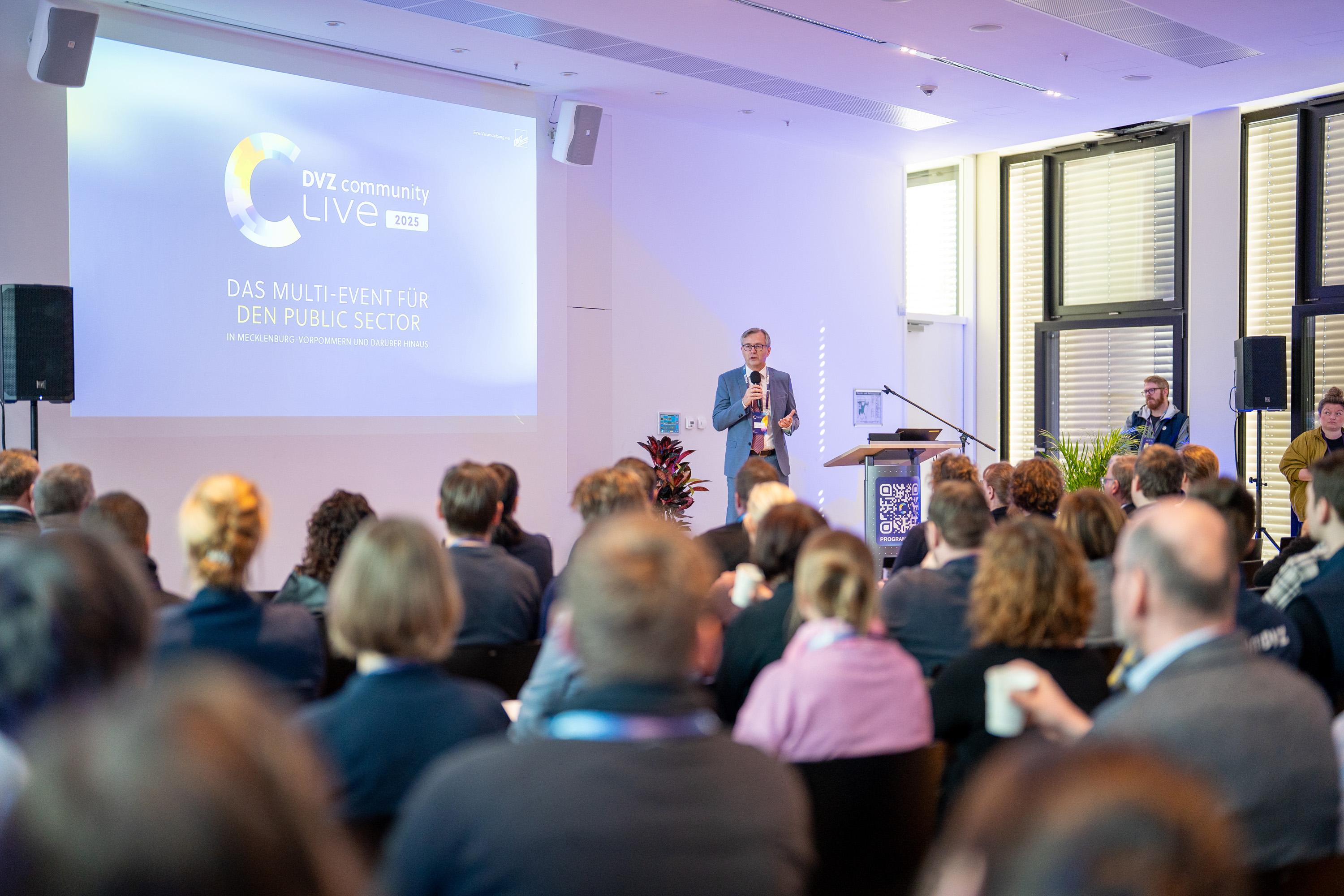 Finanzminister Dr. Heiko Geue steht auf der Bühne im Raum Mecklenburg. Im Hintergrund ist eine Präsentation mit dem Titel "DAS MULTI-EVENT FÜR DEN PUBLIC SECTOR" zu sehen.