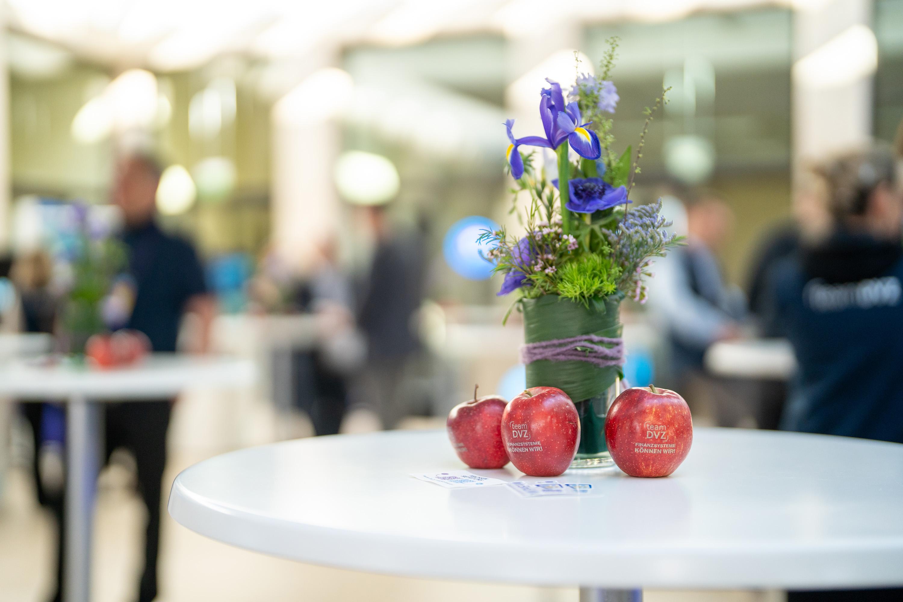 Ein Tisch mit drei roten Äpfeln und einer Blumenvase, umgeben von unscharfen Personen im Hintergrund.