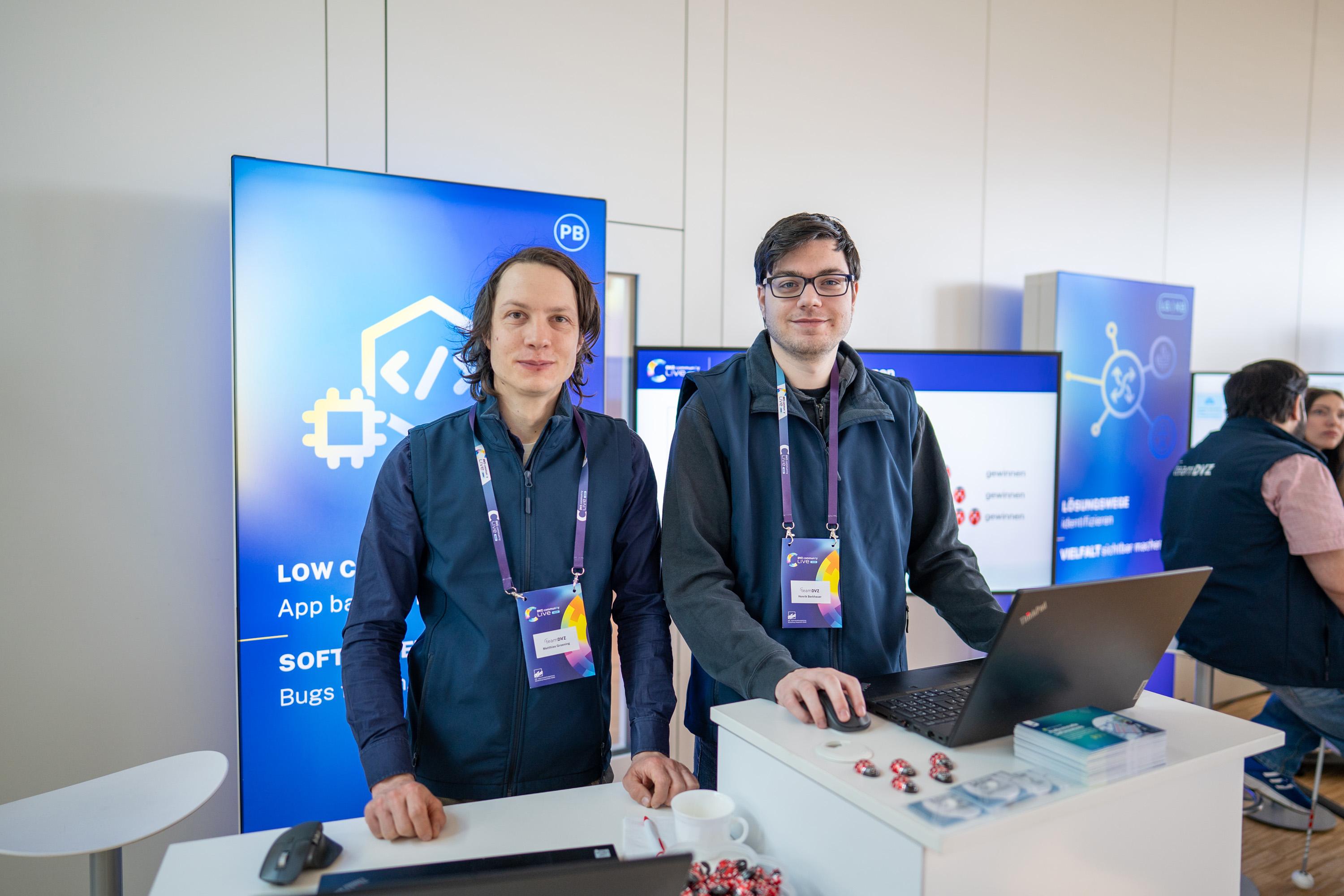 Zwei Männer stehen an einem Messestand mit Laptops. Im Hintergrund sind Banner mit Software-Themen sichtbar.