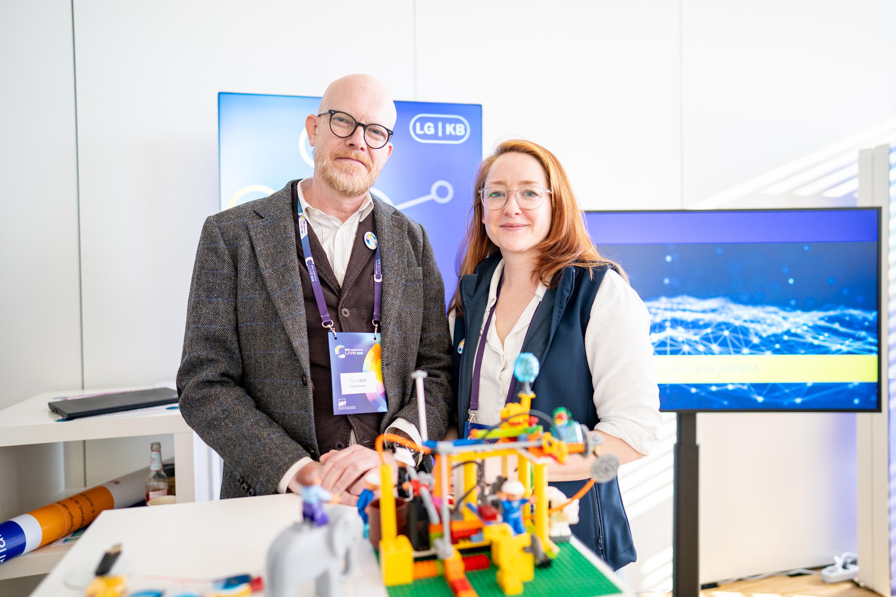Zwei Personen stehen an einem Messestand. Der Mann trägt eine Brille und einen grauen Anzug, die Frau hat lange rote Haare und eine blaue Weste. Vor ihnen steht ein LEGO-Modell.
