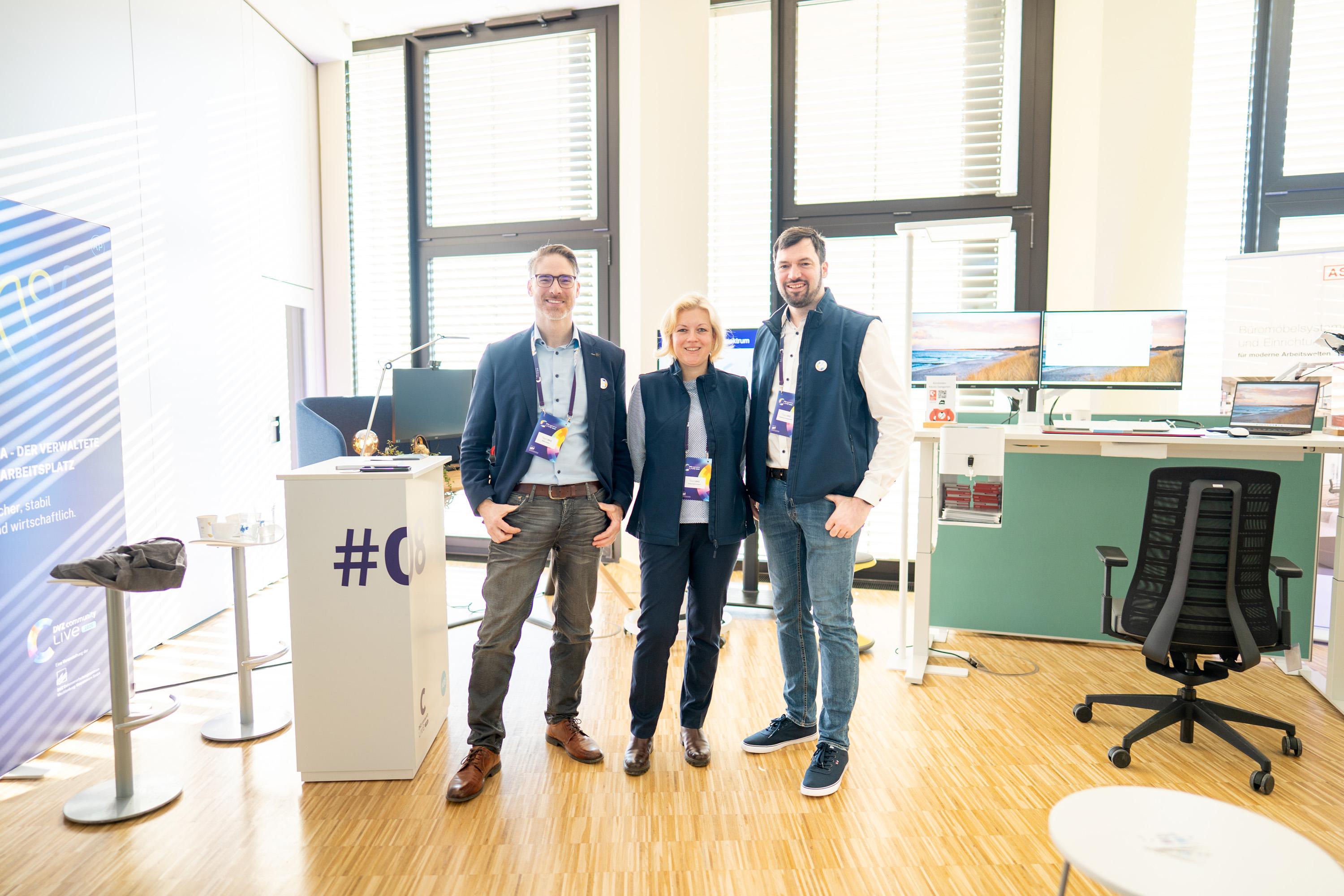 Drei Personen stehen in einem modernen Büro-Setup auf einer Messe. Sie tragen Business-Kleidung und lächeln. Im Hintergrund sind Computer und ein Messestand sichtbar.