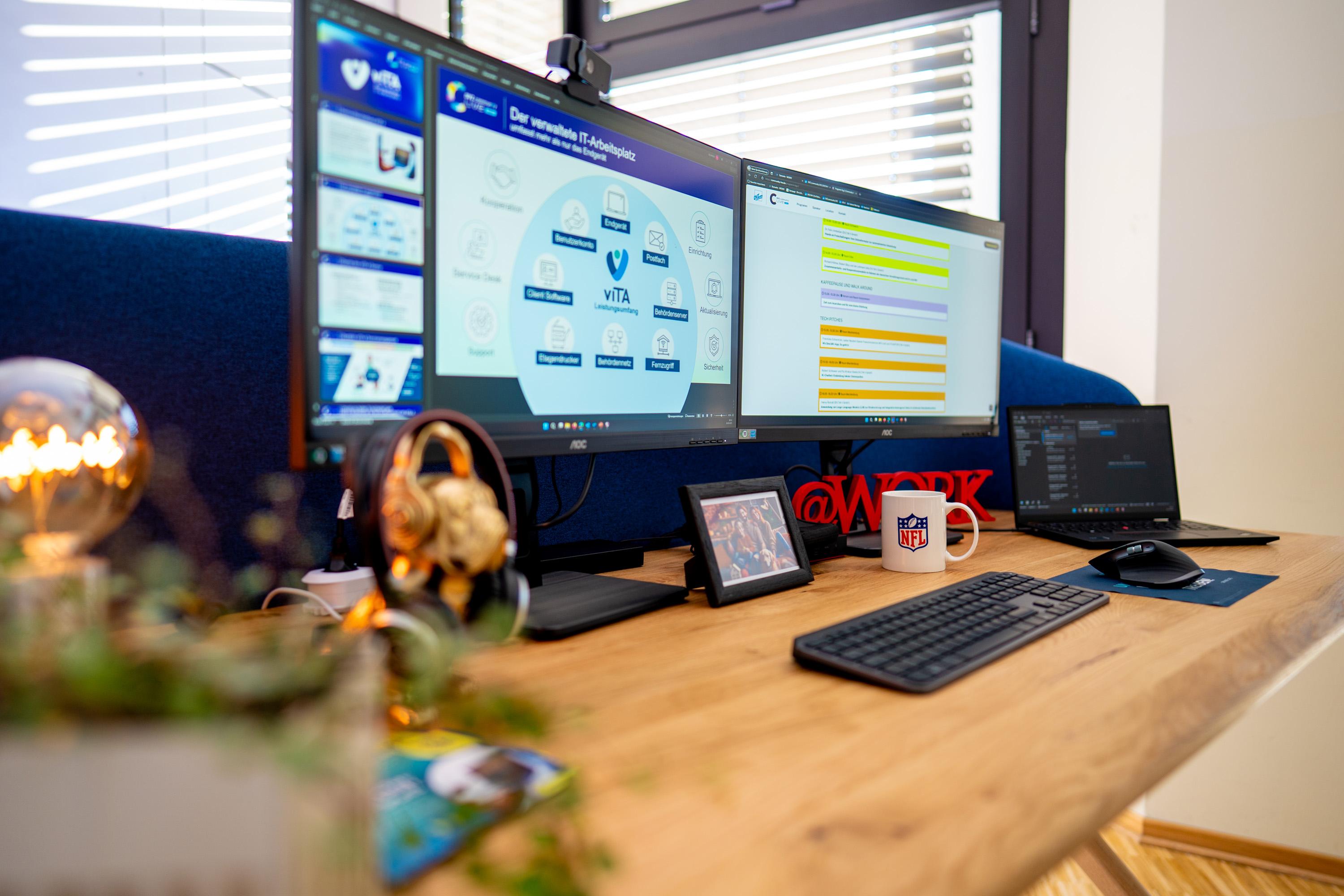 Büroarbeitsplatz mit zwei Monitoren, Laptop, Kaffeetasse und Pflanzen. Auf einem Monitor ist eine Präsentation zu sehen.