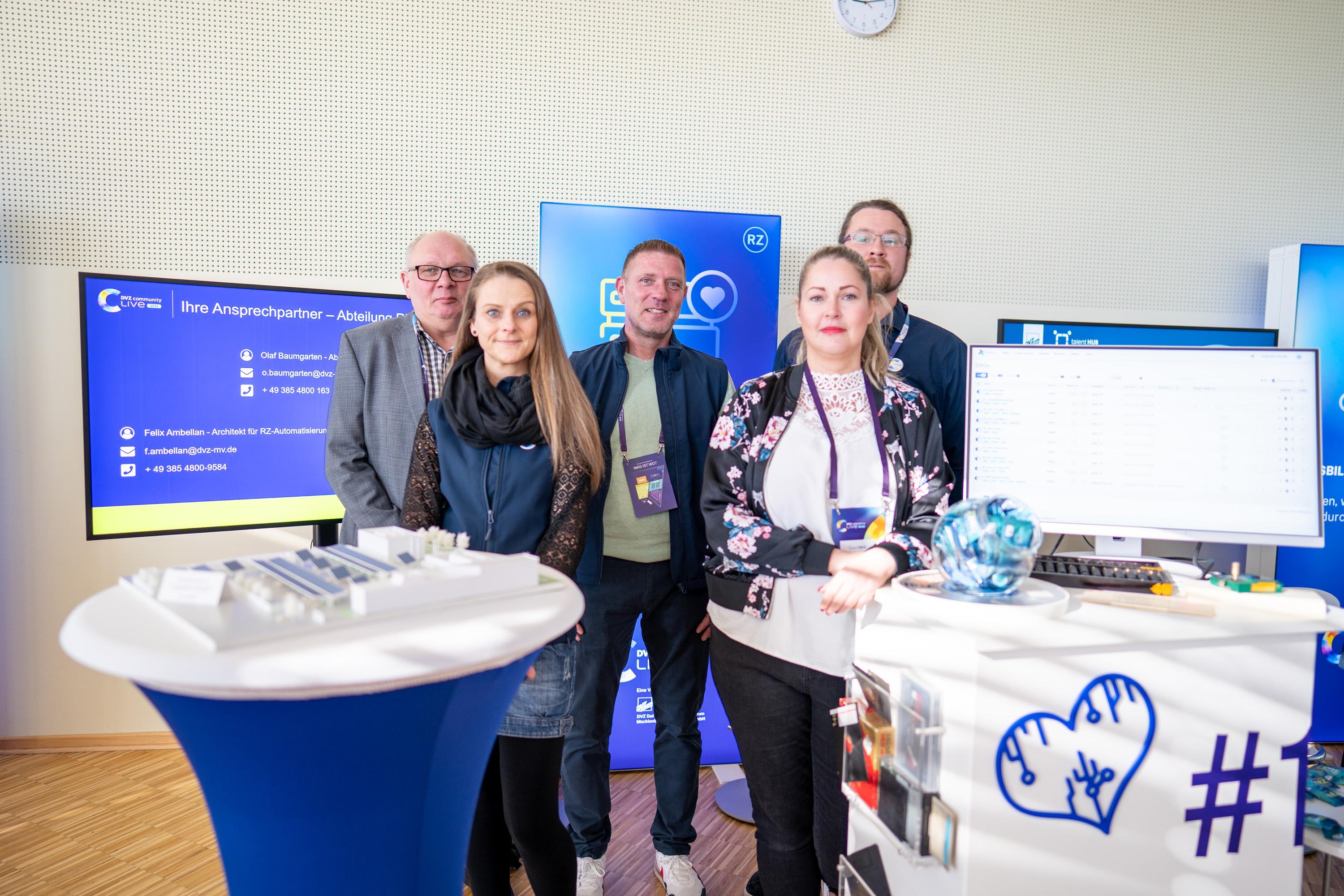 Gruppe von fünf Personen an einem Messestand, umgeben von Informationsmaterialien und einem Computer. Die Personen stehen lächelnd zusammen, einige tragen Namensschilder.