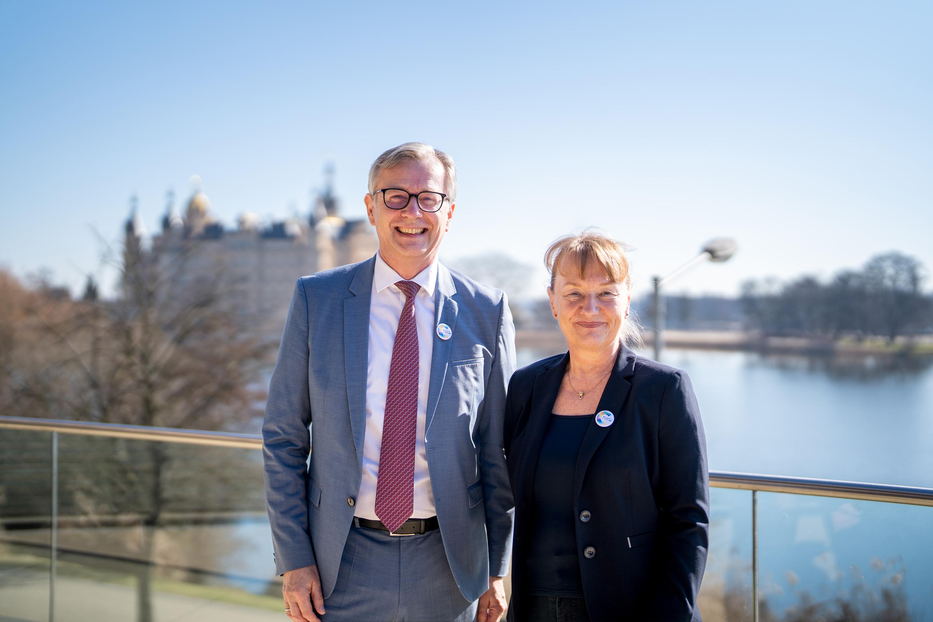 Finanzminister Dr. Heiko Geue und Uta Knöchel stehen lächelnd auf der Dachterasse der IHK zu Schwerin, im Hintergrund das Schweriner Schloss.