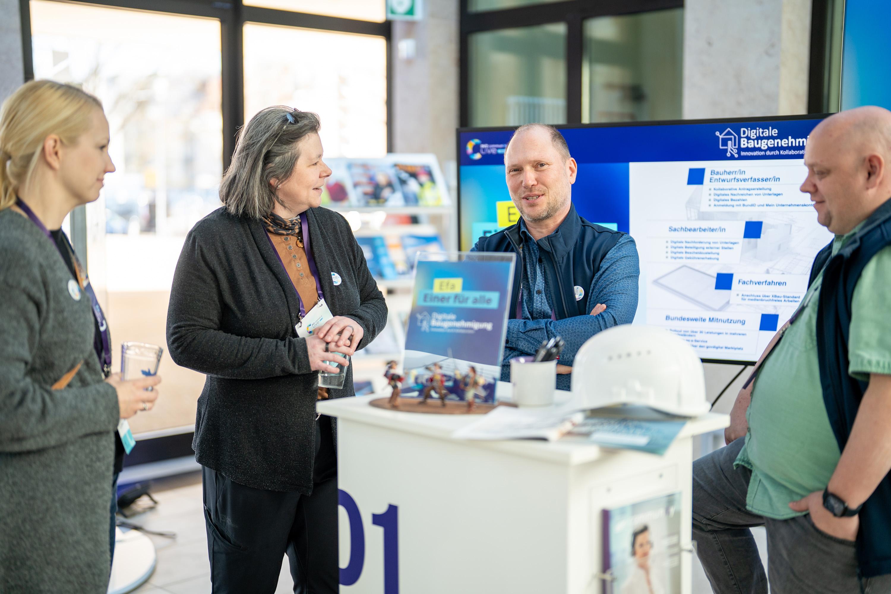 Vier Personen stehen an einem Messestand und diskutieren. Im Hintergrund ein Bildschirm mit Informationen zur digitalen Baugenehmigung.