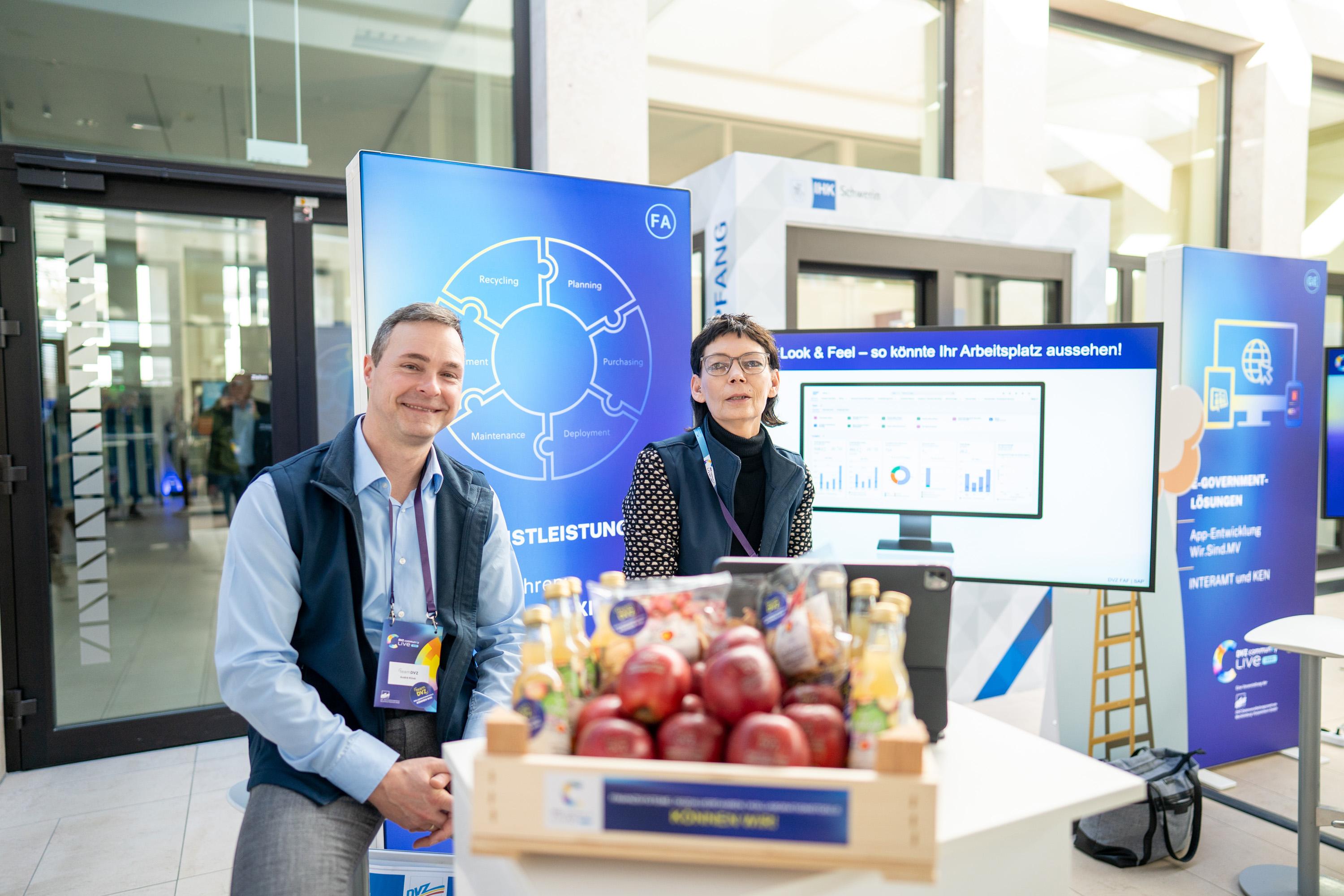 Zwei Personen an einem Messestand, umgeben von Obst und Getränken, mit einem Bildschirm im Hintergrund, der Diagramme zeigt.